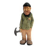 Hand-carved wooden figurine titled "Johannes with Shovel" by Bill Erickson, depicting a bearded man in a green shirt and cap, skillfully handling a pickaxe.