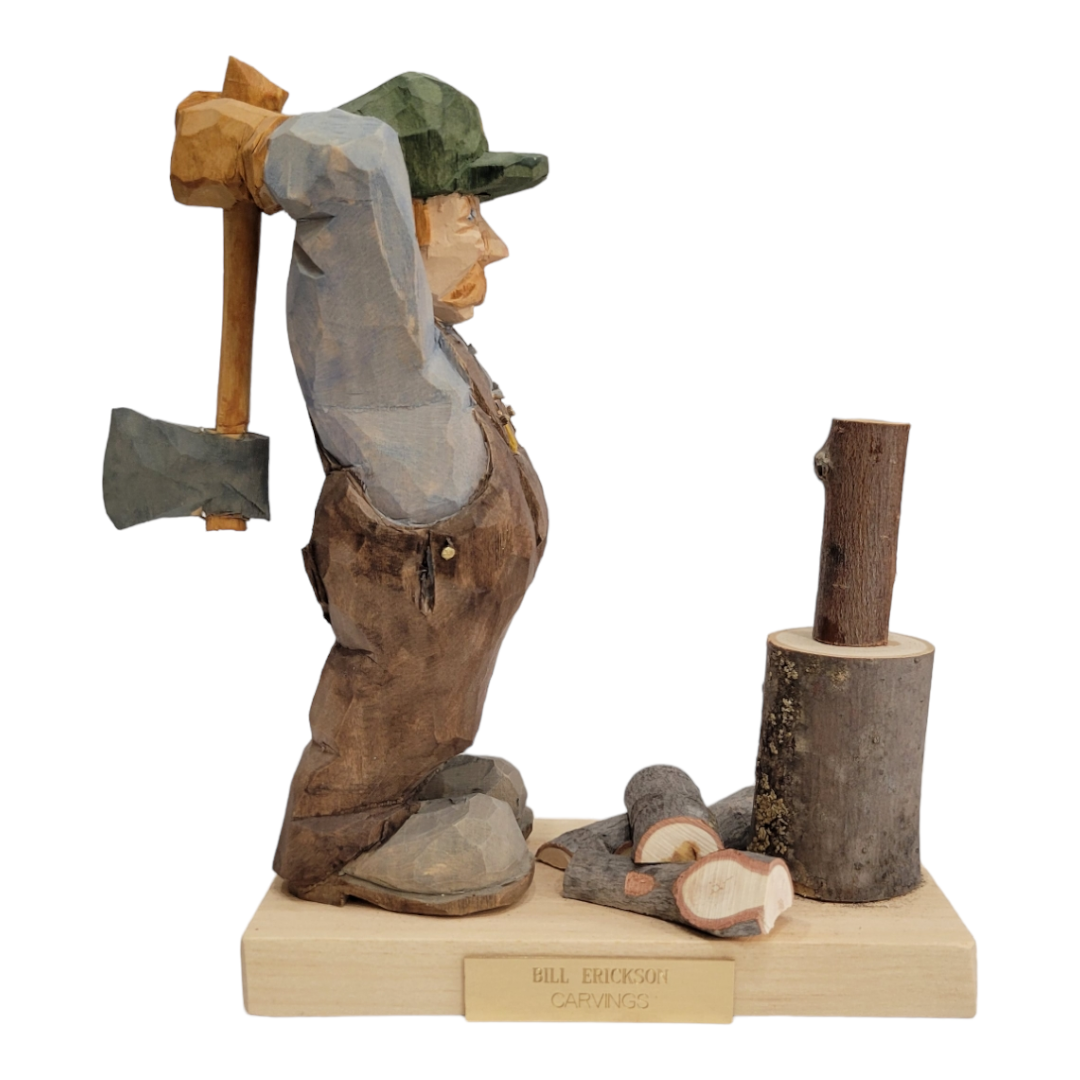 Figurine: 'Woodchopper' by Bill Erickson (Copy) features a meticulously hand-painted wood carved figure of a man in overalls, poised to swing an axe at a log resting on a tree stump. The scene is completed with several pieces of chopped wood on the base and is labeled with Bill Erickson Carvings.