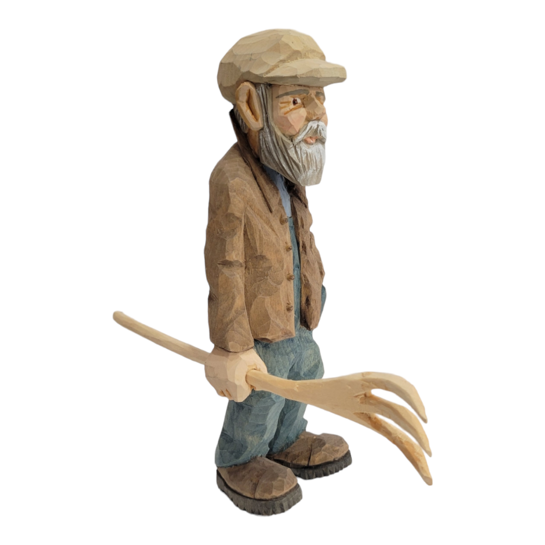 This wood-carved figurine, titled "Johannes" by Bill Erickson, depicts a bearded man wearing a cap and coat while holding a pitchfork.