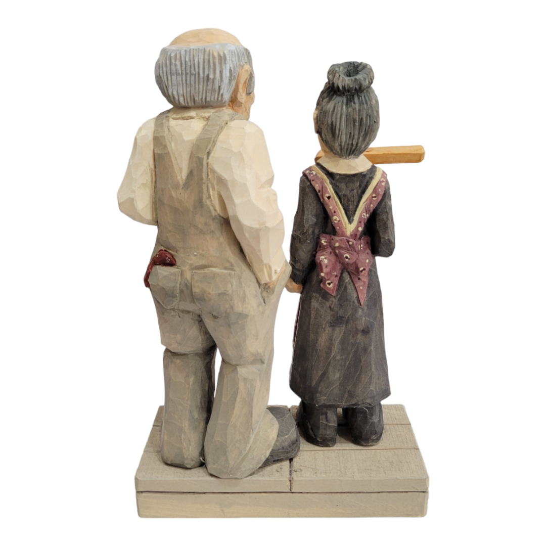 Standing at 7 inches tall, the "Salt of the Earth" wooden figurine by Bill Erickson features an elderly couple elegantly poised on a platform. The man is dressed in suspenders, while the woman wears her hair in a bun and dons an apron, both captured in painted attire that embodies their humble essence.