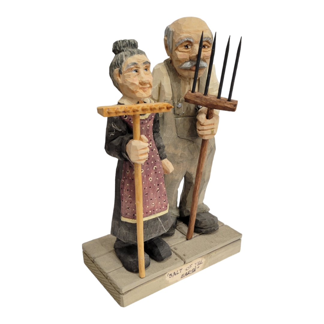 The "Salt of the Earth" figurine by Bill Erickson is a beautifully crafted 7-inch tall wooden sculpture that captures the essence of an elderly couple holding farming tools. The woman wears a dress, and the man is dressed in overalls, showcasing their timeless presence as wood-carved figures.