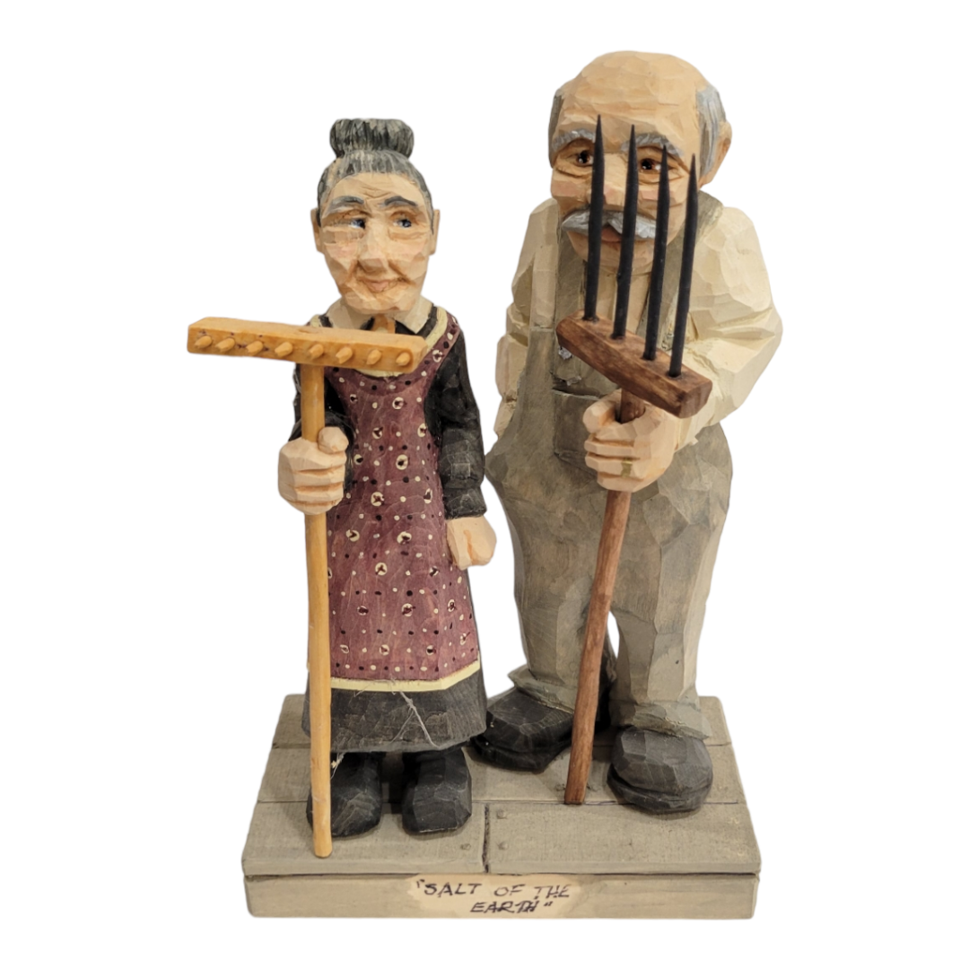 Standing at 7 inches tall, the "Salt of the Earth" figurines by Bill Erickson depict two intricately carved wood figures—a woman with a rake and a man with a pitchfork—that beautifully embody rural life. Situated on a base bearing their title, their attire embodies the enduring spirit of farming.