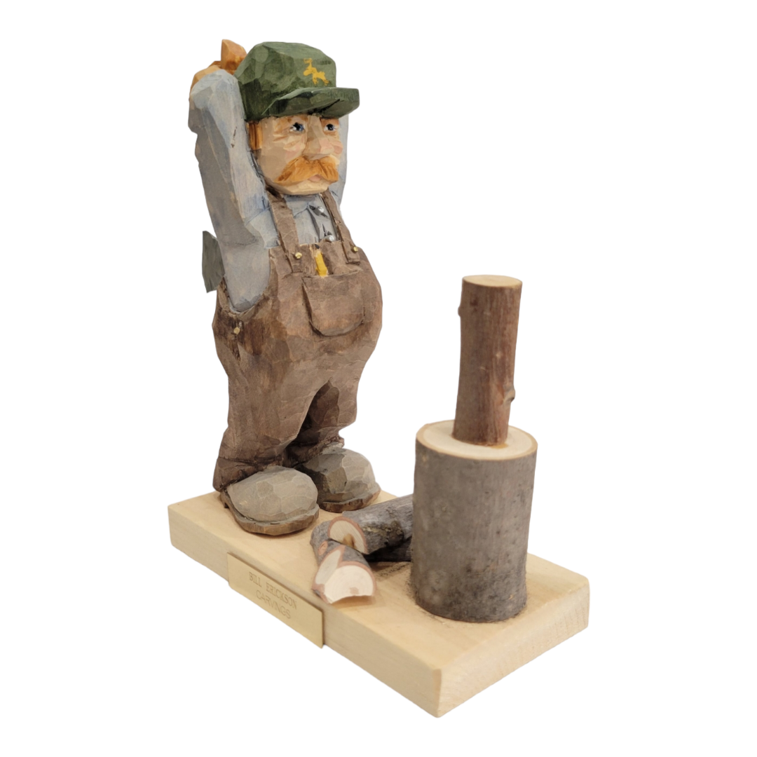 The Woodchopper figurine by Bill Erickson depicts a man in overalls and cap, axe poised behind his head, standing beside a log on a base with a small plaque.