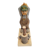 The Woodchopper by Bill Erickson is a hand-carved wooden figurine depicting a man in a green hat and overalls, leaning on an axe beside a log, elegantly poised on a wooden base.