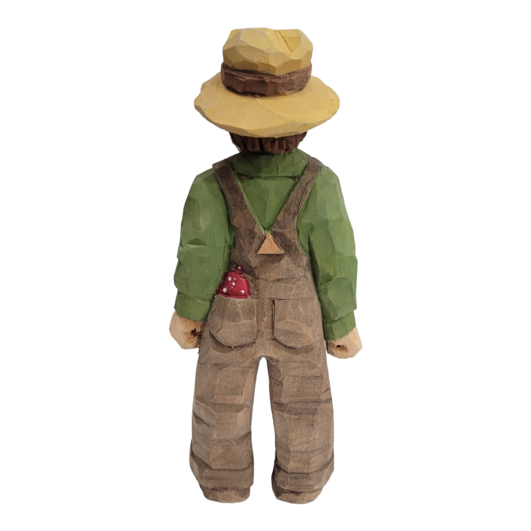 The "Happy Farmer" figurine by Bill Erickson is a 7-inch wood carved figure depicting a person in a green shirt, brown overalls, and straw hat, viewed from behind.