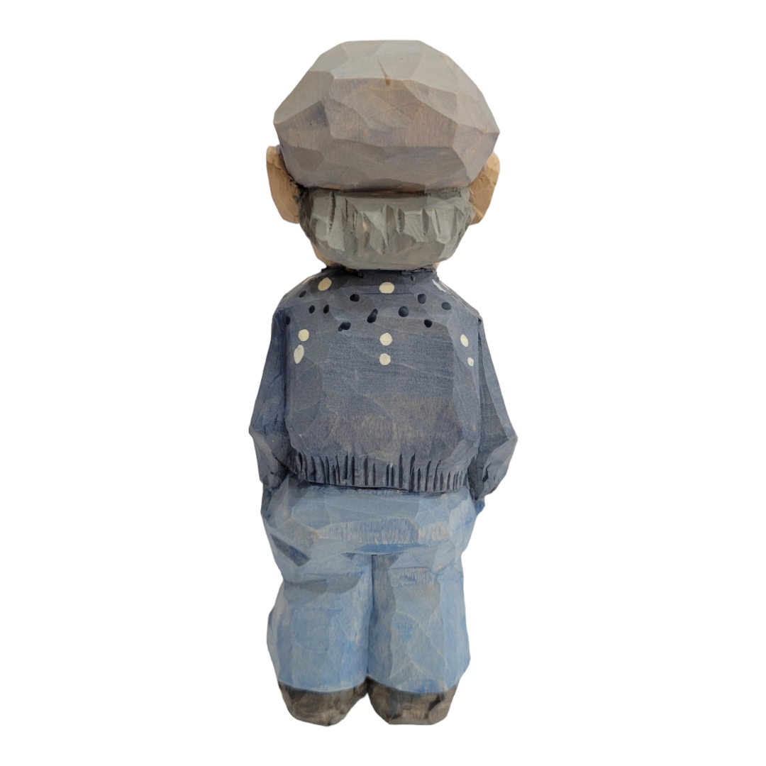 The "Lars" figurine by Bill Erickson is a 6-inch wood carved figure that portrays a person viewed from the back, wearing a cap and dressed in blue clothing with white dots on the jacket.