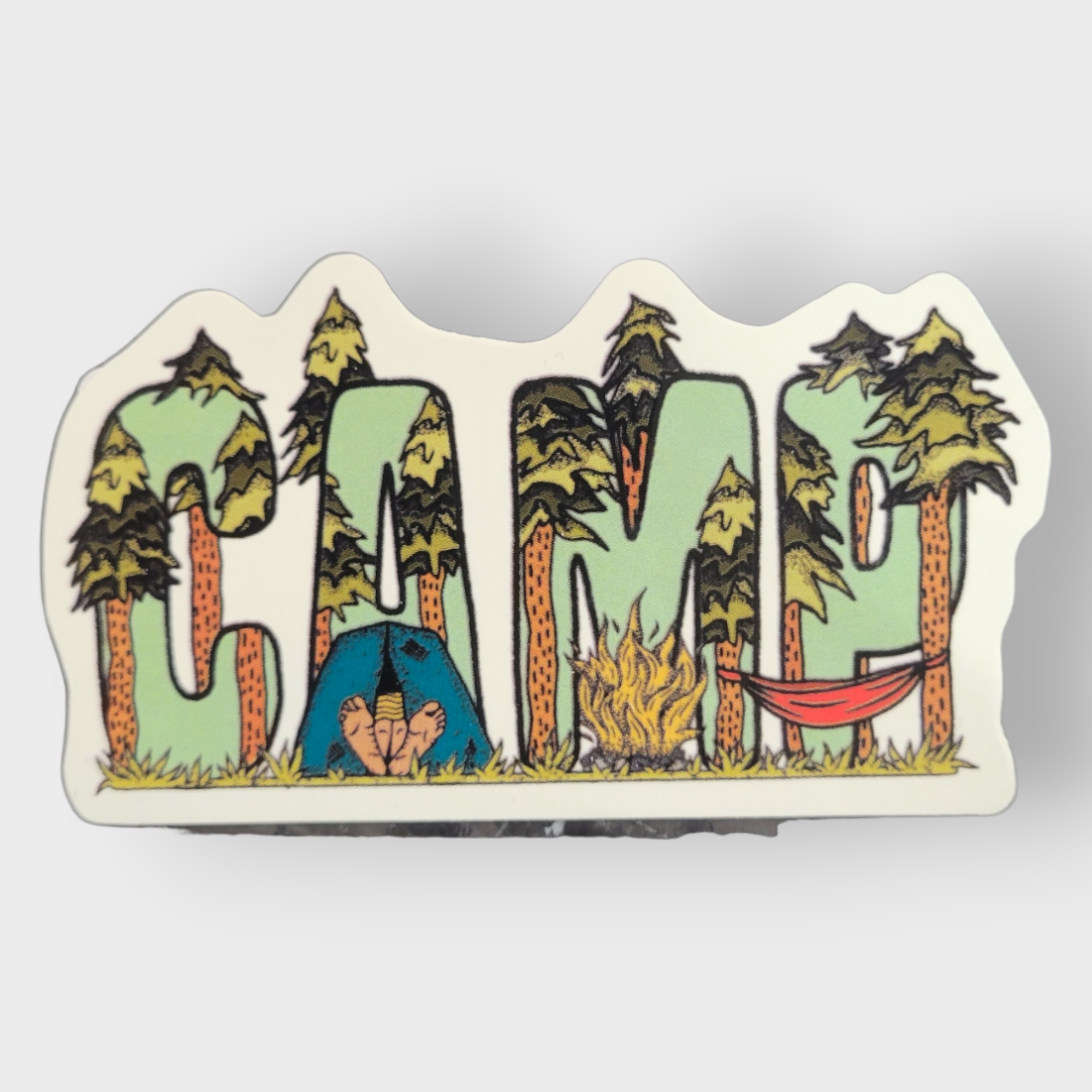 The Camping Hiking Sticker with Hammock showcases the word CAMP illustrated with letters creatively shaped from trees, along with a tent, campfire, and hammock intricately woven into the design to embody the spirit of adventure.