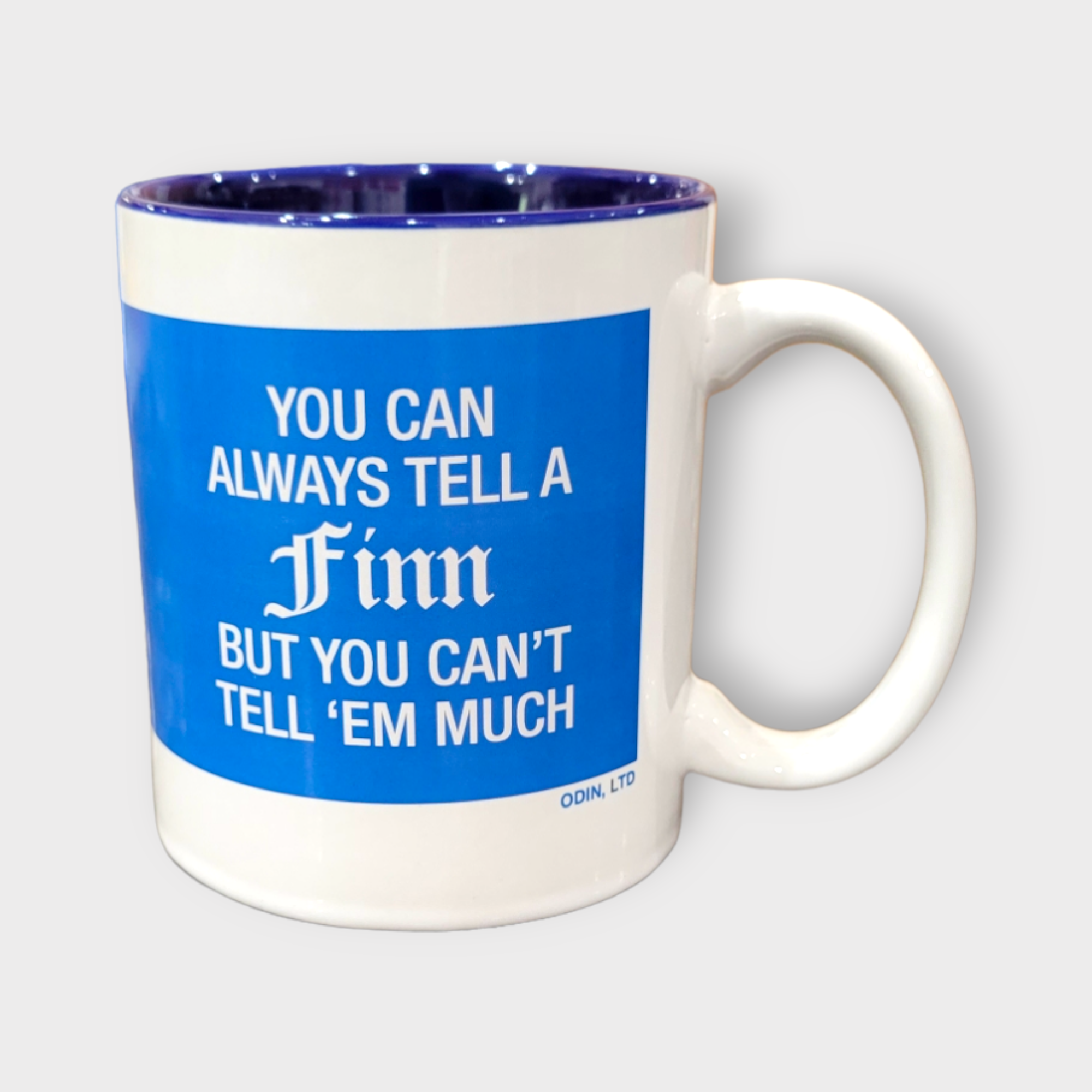 This 11 oz "Tell A Finn" mug features a white ceramic exterior with a blue interior and humorously states: "You can always tell a Finn, but you can't tell 'em much.