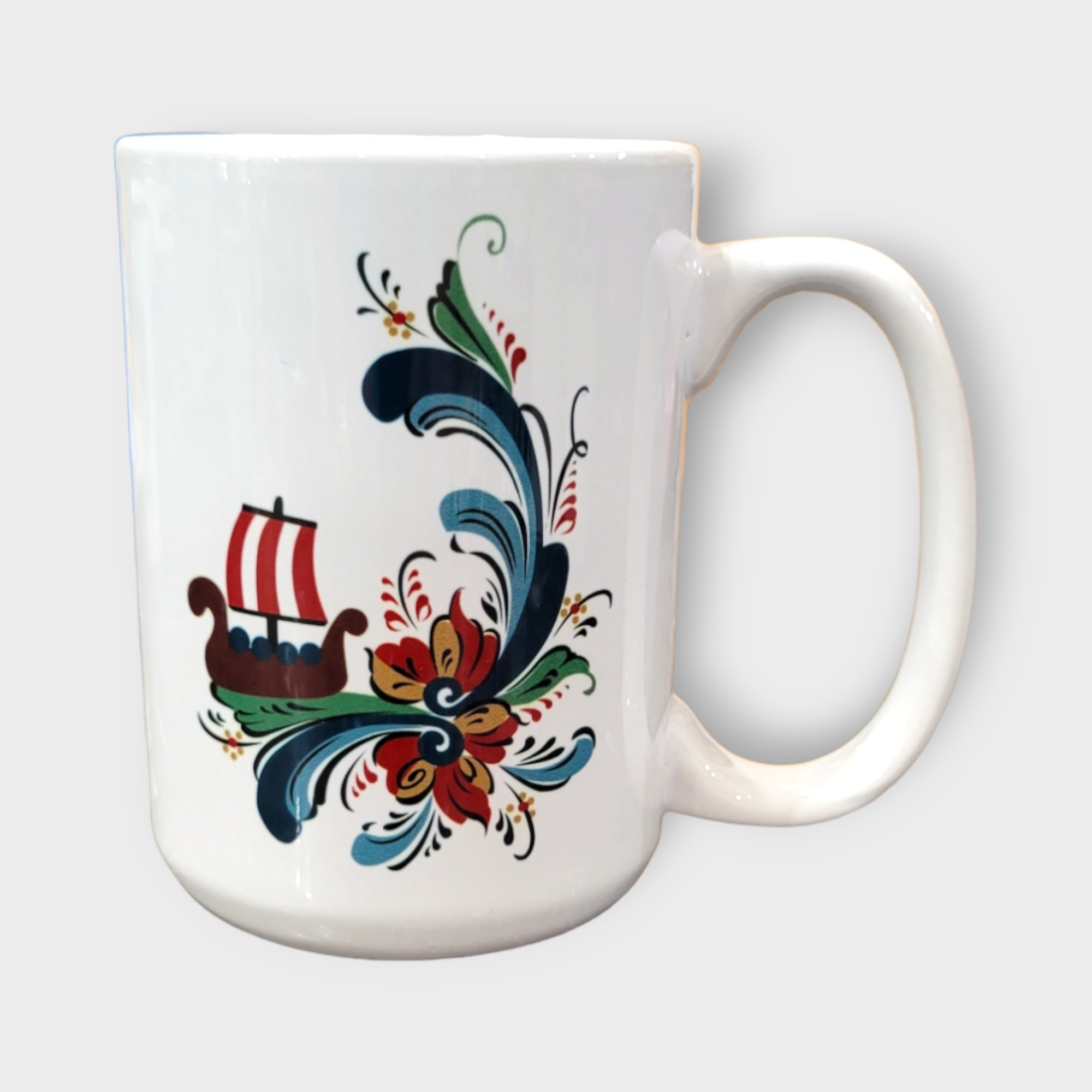 Mug: Viking Ship on Rosemaling (15oz) – a white ceramic mug featuring a red and white Viking ship and colorful Scandinavian folk art floral designs, photographed on a plain light background.