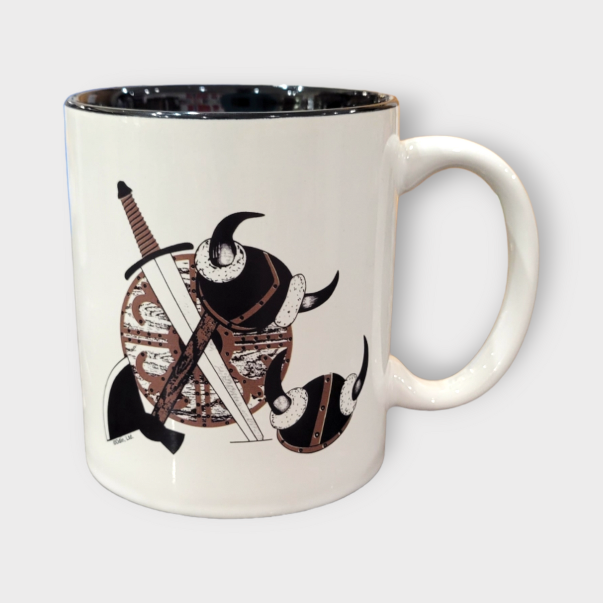 The Coffee Mug: Viking Coat of Arms (11oz) is a white ceramic mug with a brown and black illustrated Viking helmet, sword, and shield design. Dishwasher safe.