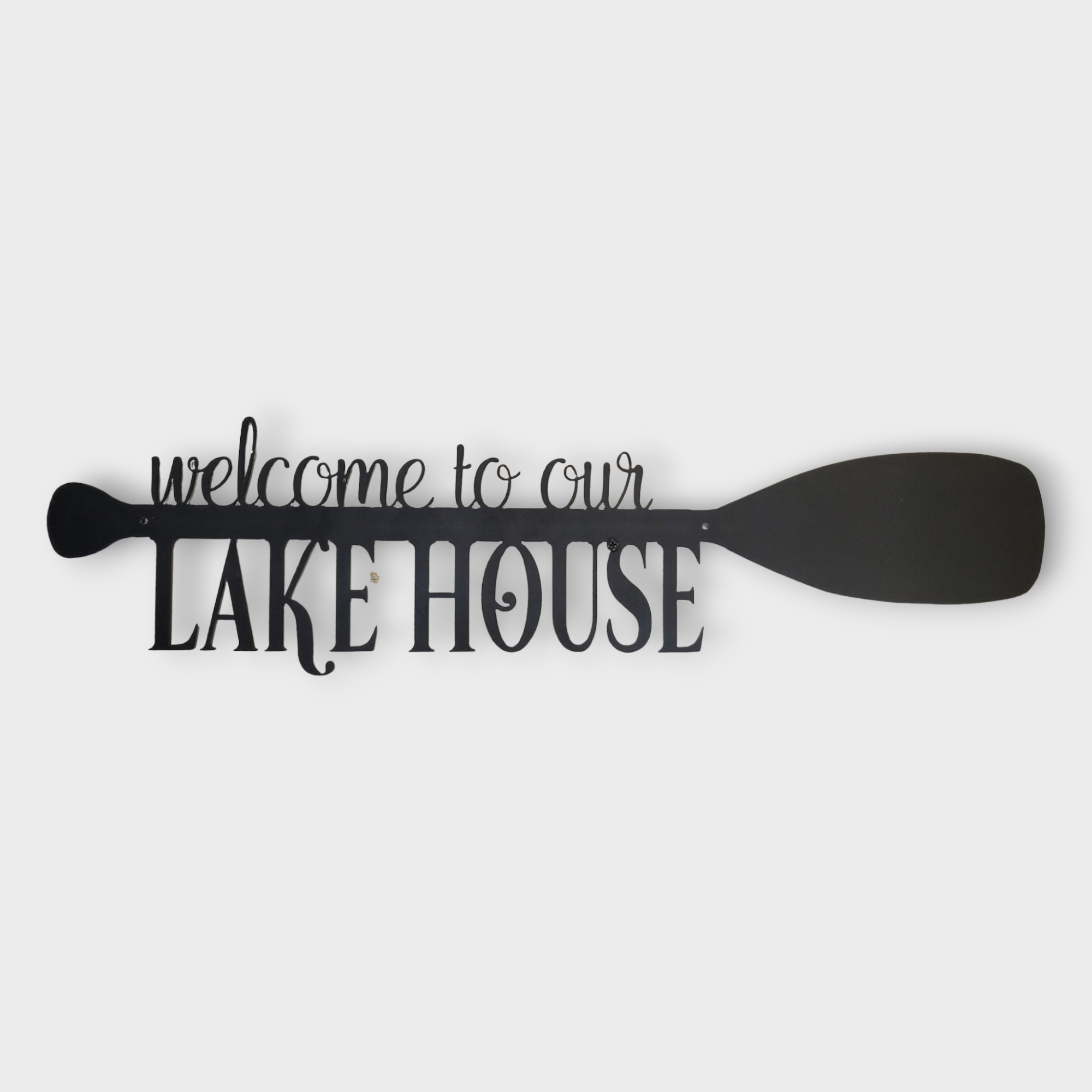 The "Wall Decor: Welcome to Our Lake House Paddle, Metal" is an elegant piece of black metal wall art that spells out "Welcome to our Lake House" and includes a paddle-shaped accent on the right side.