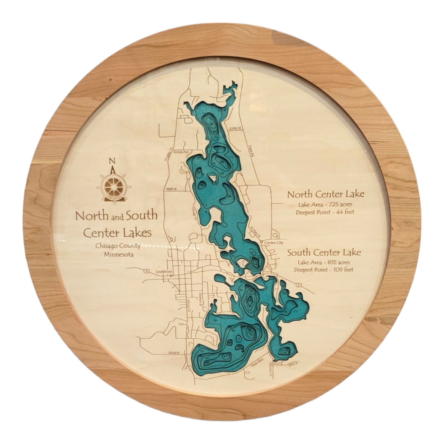 The Lake Map: Round Map North & South Center Lakes is wooden wall art showcasing a layered map of the lakes in Chisago County, MN. Made from natural wood, it highlights lake areas and depths with a compass rose at the top left for personalization.