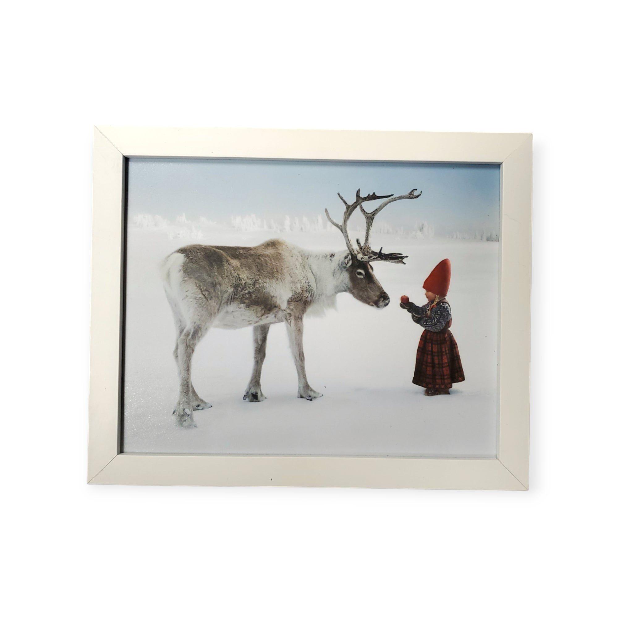 A child in a red hat and winter clothing stands in the snow, reaching out to a reindeer, perfectly depicting the enchanting scene titled "Artwork: Anja with Reindeer - Christmas Wish." This beautifully captured moment set against a snowy landscape is available as a framed print.