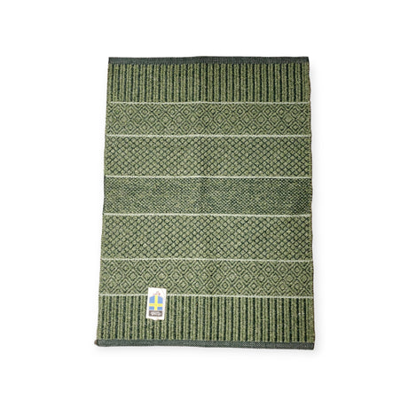 The Rug: Mixed Alice - Olive (Cotton & Vinyl) is showcased on a plain background, displaying its green patterned design with various textured stripes reminiscent of a Swedish vinyl woven rug. A small tag at the bottom corner emphasizes its durable, skid-resistant construction, perfect for versatile indoor and outdoor use.