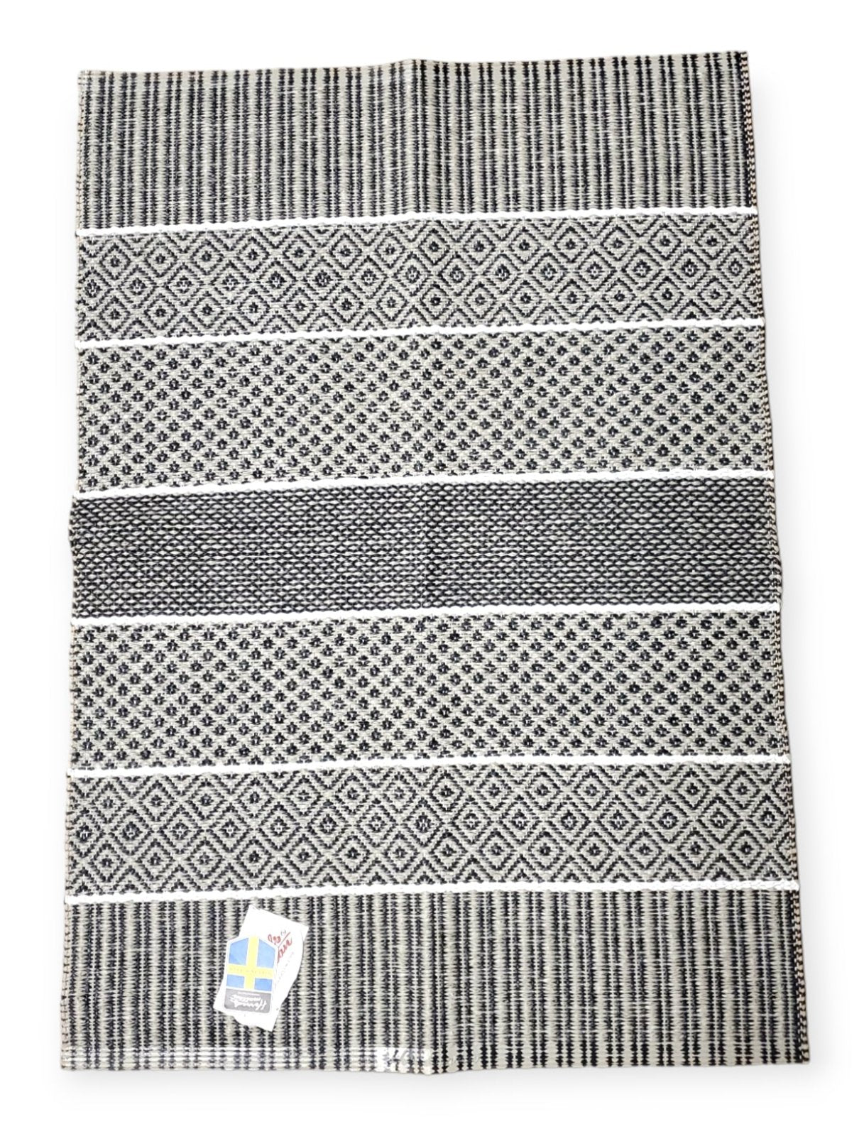 Presenting the "Rug: Alfie Graphite," a Swedish vinyl rug in black and white, showcasing geometric patterns. It comes with a non-skid backing and features a durable square label located in the bottom left corner.