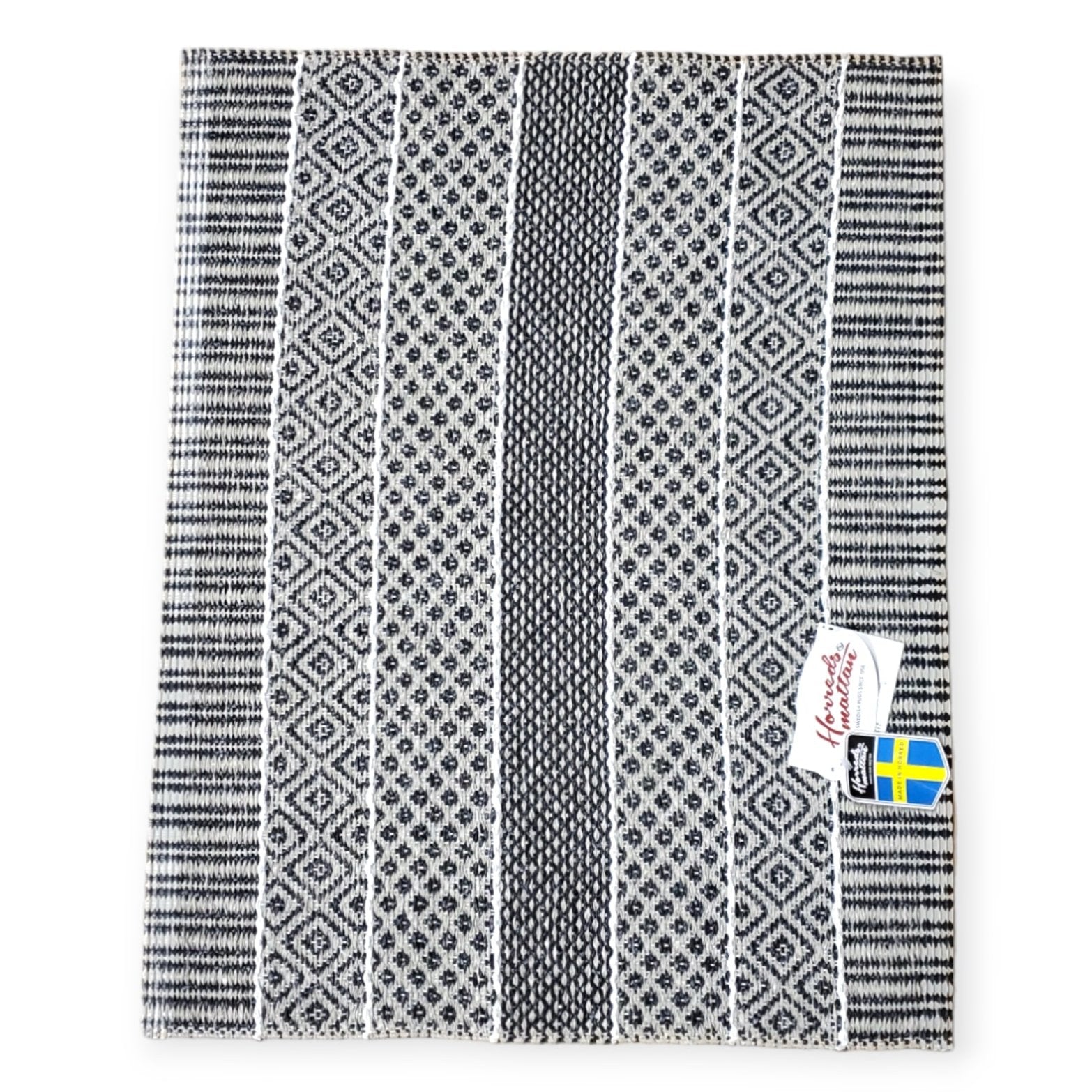 The "Vinyl Rug: Horredsmattan - Alfie - Graphite" features a black and white pattern reminiscent of a woven vinyl rug, showcasing intricate designs along with brand and Swedish flag tags.