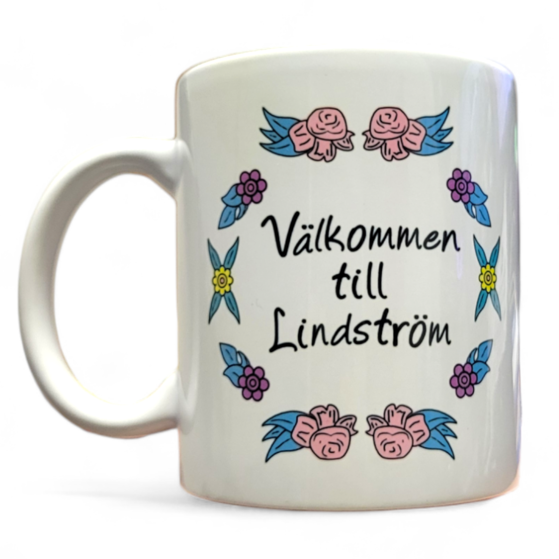 The "Välkommen till Lindstrom" mug is a white ceramic 11oz cup adorned with the words "Välkommen till Lindström," accompanied by a floral motif of pink roses and blue and purple flowers. It is dishwasher safe, making it ideal for everyday use.