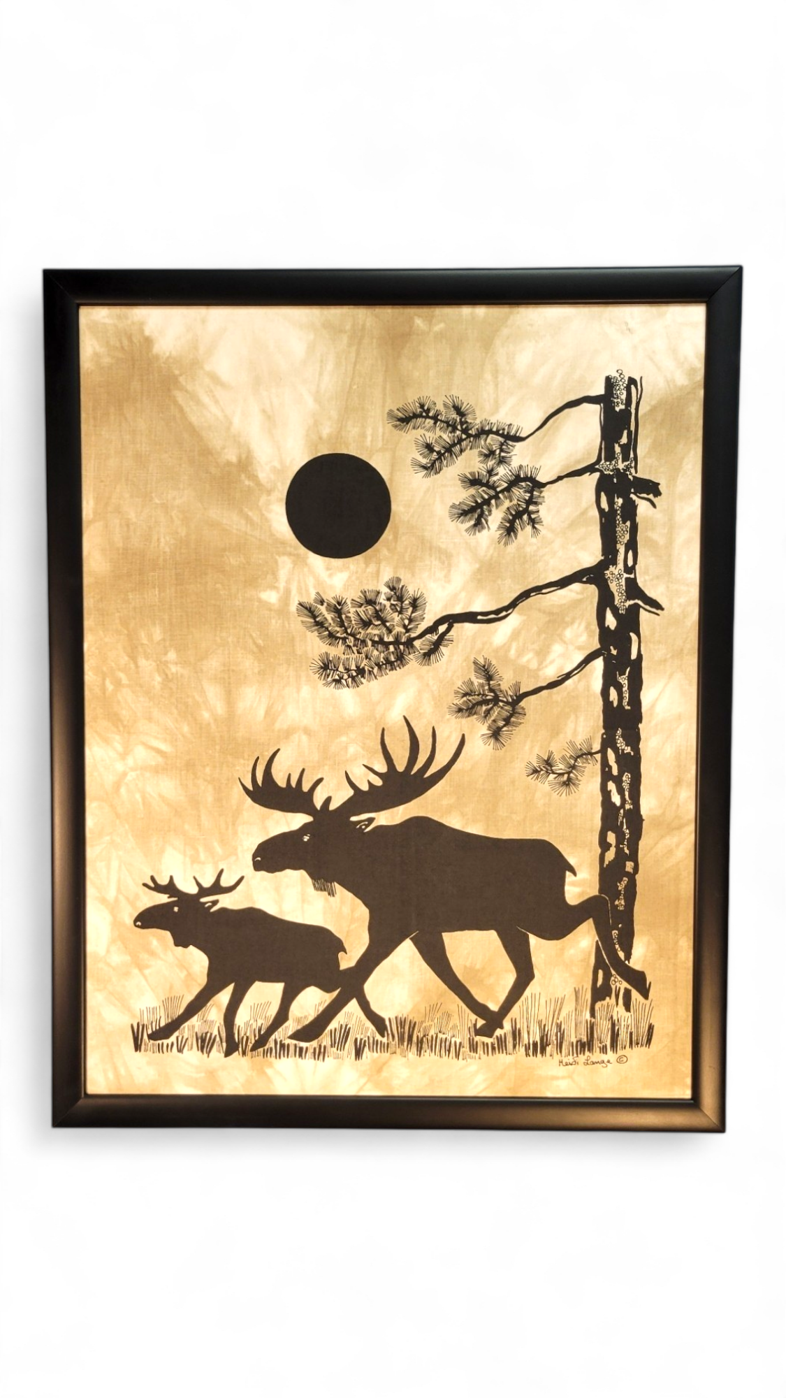 This stunning framed silkscreen by Heidi Lange, titled "Under the Pine Tree," beautifully depicts two moose gracefully walking in front of a tree beneath a round sun, with dimensions of 19.5x24.5 inches.