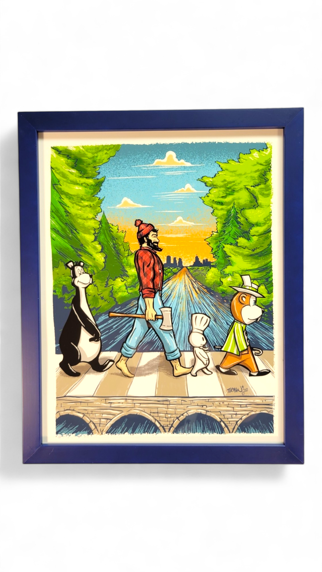 Framed screen print illustration titled "Abbey Road 11x14" by Adam Turman, features four cartoon characters walking on a crosswalk amidst a vibrant landscape with green trees and a blue sky, capturing Turman's iconic Minnesota style.