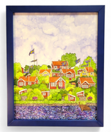 Artwork titled "Brändaholm" by Greta Lann, featuring a framed painting of a scenic village adorned with red and white houses, Swedish flags, lush greenery capturing the essence of Karlskrona's Brändaholm charm, and a calm body of water in the foreground.