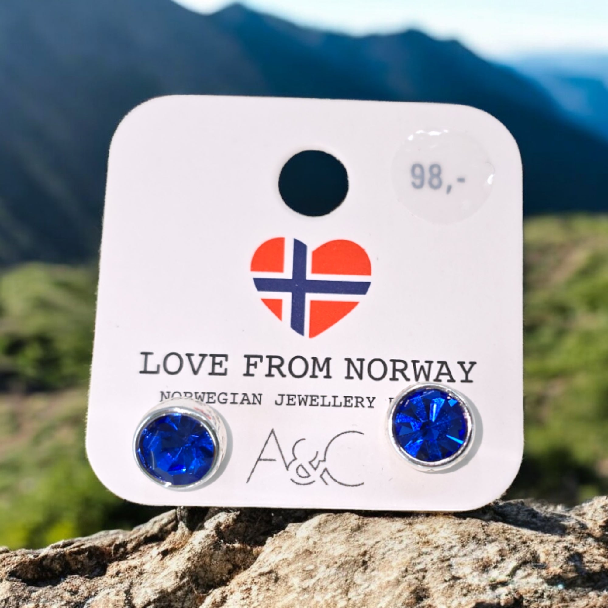 Sapphire crystal earrings with silver posts from A&C Norway, presented on a card featuring a Norwegian flag heart logo and labeled Love from Norway. These earrings elegantly capture the spirit of Norwegian nature, with a backdrop of blurred mountainous landscape.