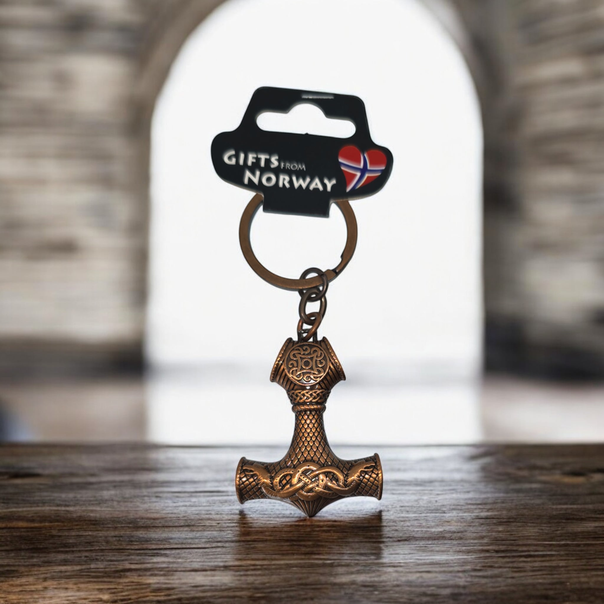 A Key Chain: Thor's Hammer Bronze showcasing detailed Norse designs is presented on a wooden platform, labeled Gifts from Norway, with a Norwegian flag in the background.