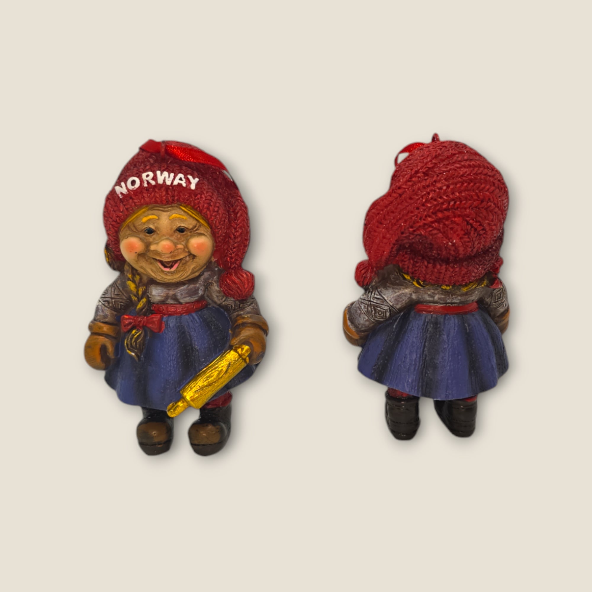 Front and back views of the smiling "Figurine: Nisse Wife with Rolling Pin 3.5"" wearing a red hat that reads Norway, dressed in a blue outfit, and holding a yellow rolling pin.