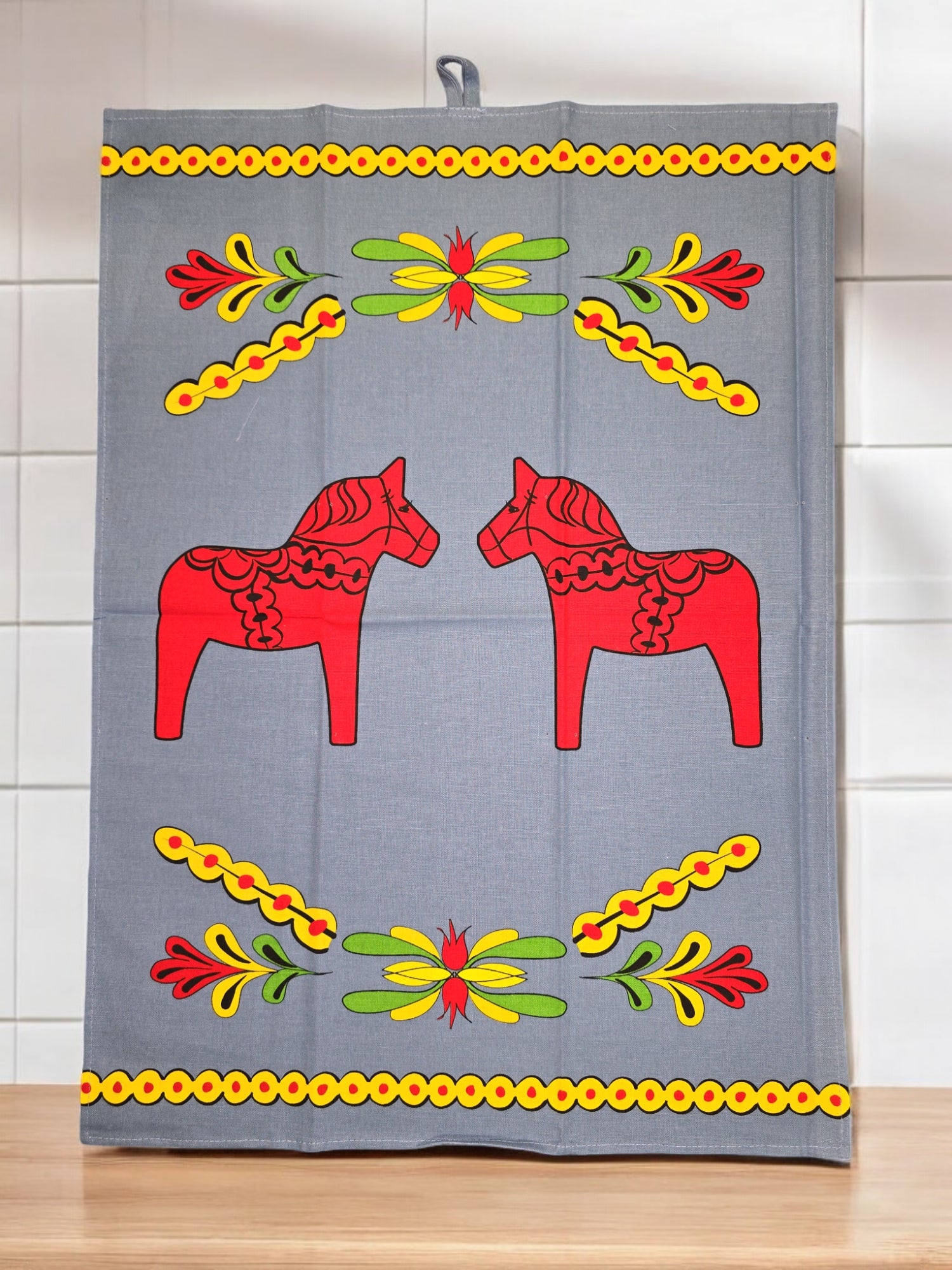 The tea towel, featuring vivid Swedish Dala Horse motifs and colorful Kurbits floral patterns, is displayed hanging against a tiled wall.