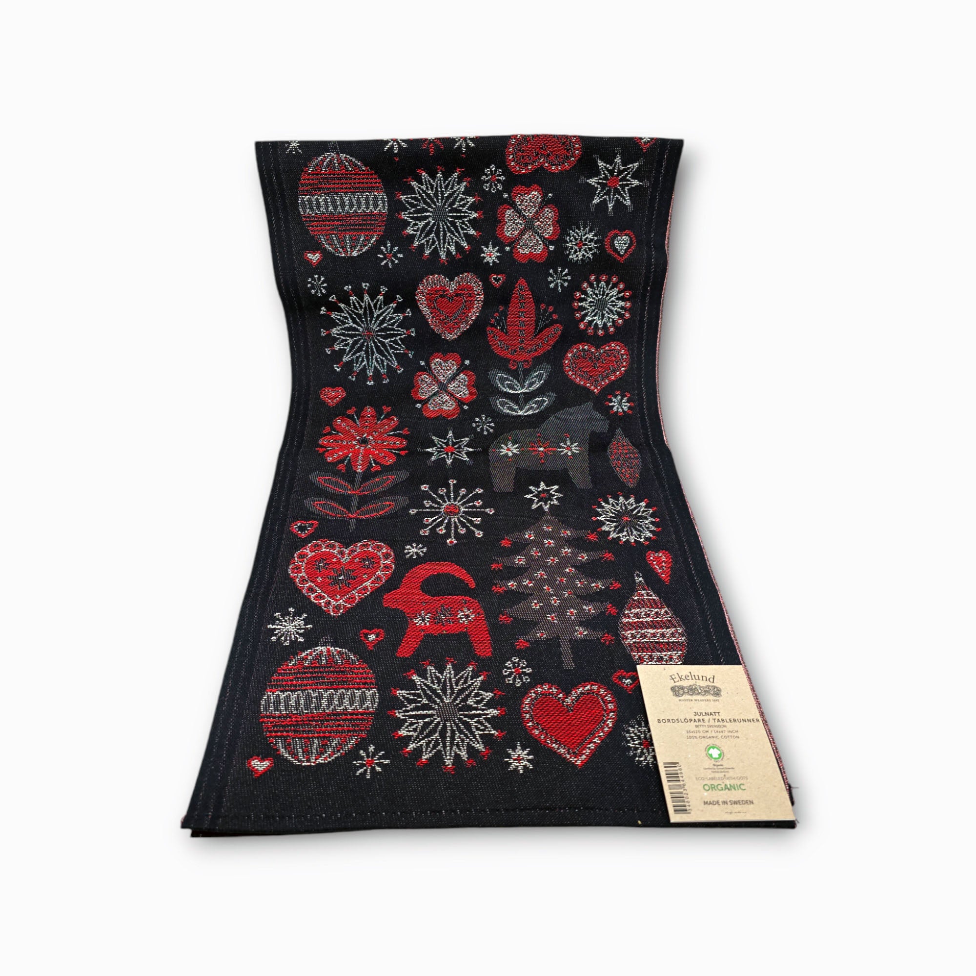 The Runner: Christmas Julnatt from Ekelund Master Weavers is a black fabric table runner adorned with festive designs of red and white hearts, snowflakes, and ornaments. Crafted from organic cotton in Sweden, it includes a tag emphasizing its organic production.