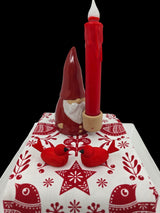 A red ceramic gnome holding a tall red candle stands on a white and red Scandinavian-patterned cloth. In front, two Bergquist Imports Wooden Scandi Birds Handcrafted Figurines from Sweden (3" long) face each other, celebrating Nordic artistry.