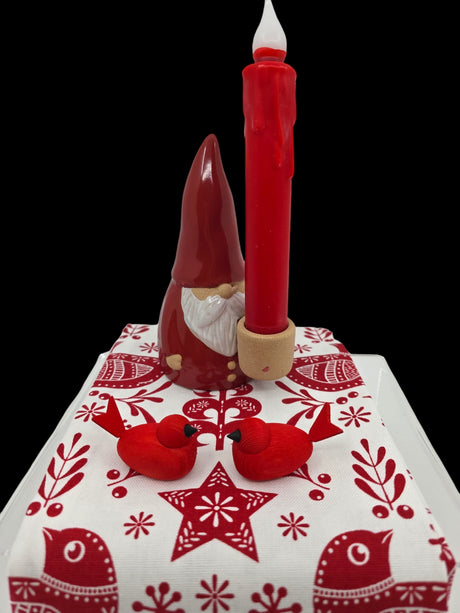 A red ceramic gnome holding a tall red candle stands on a white and red Scandinavian-patterned cloth. In front, two Bergquist Imports Wooden Scandi Birds Handcrafted Figurines from Sweden (3" long) face each other, celebrating Nordic artistry.