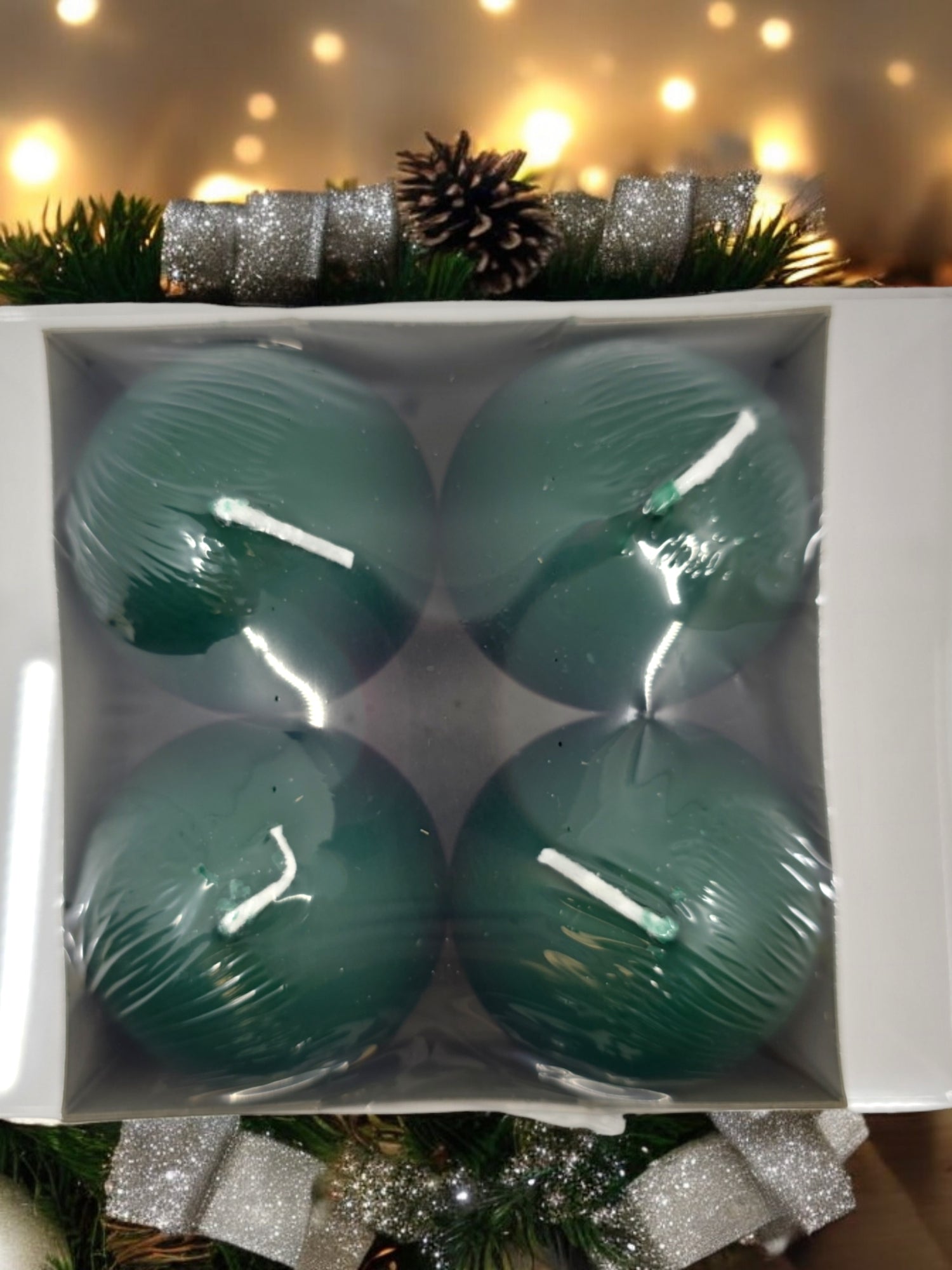 A pack of four dark green Logum Kloster Lys ball candles, placed among pine branches with ribbon and a pinecone, evokes the warm glow of Danish holiday elegance.