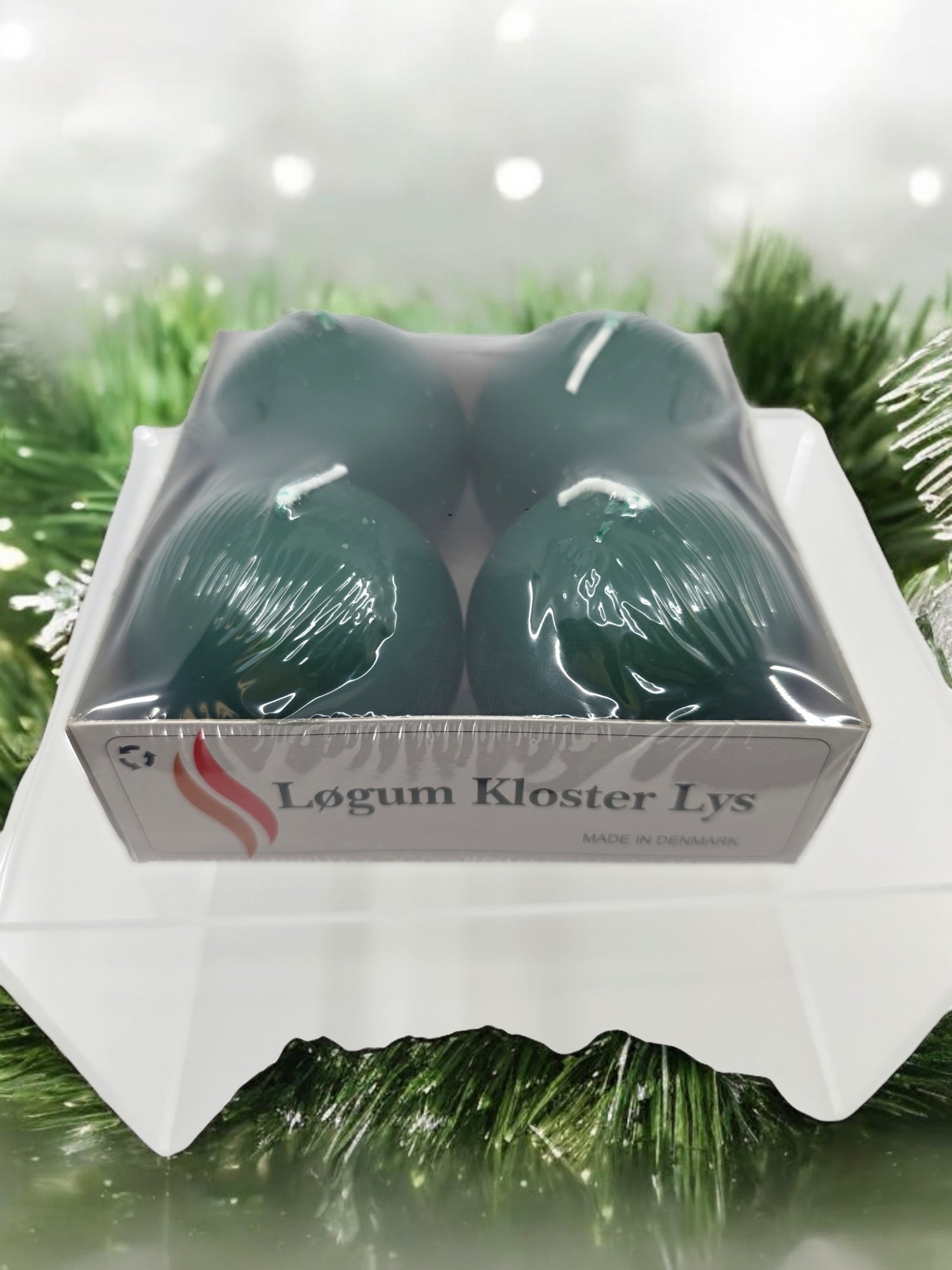 A box of four dark green 2.5" ball candles, labeled Logum Kloster Lys, is elegantly displayed on a white surface against a blurred green background.