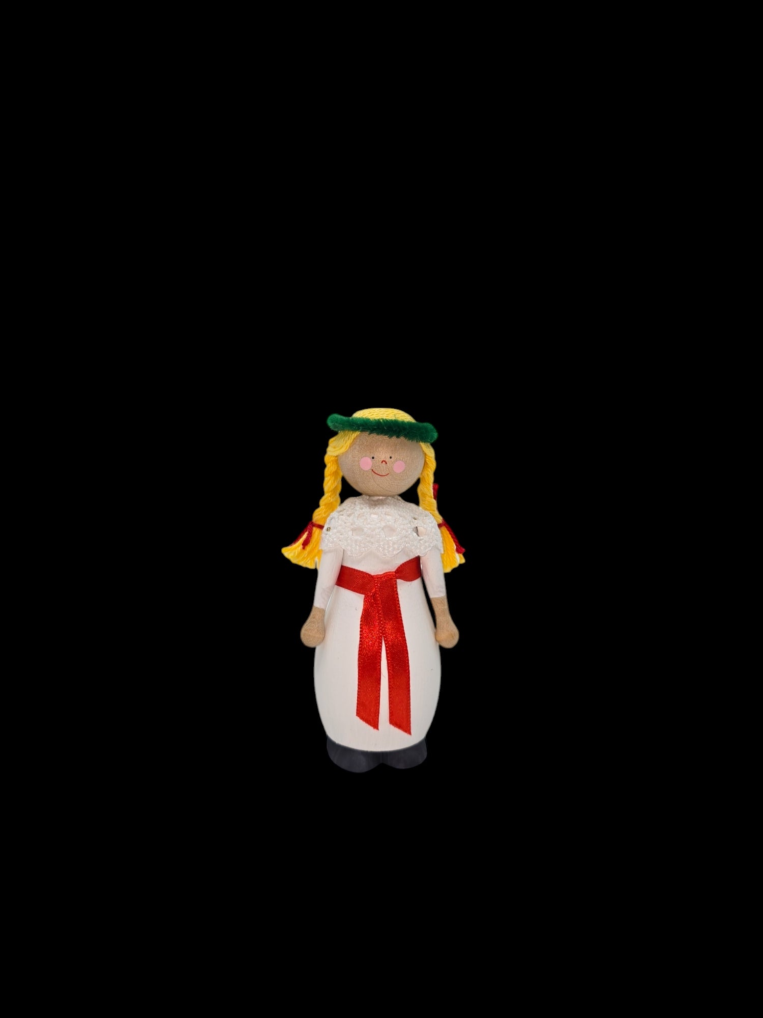 The St Lucia Hand-crafted Wood Figurine, standing 4 inches tall, features a charming portrayal with braided yellow hair and a white dress accented by a red sash and green headpiece, all set against a black background.