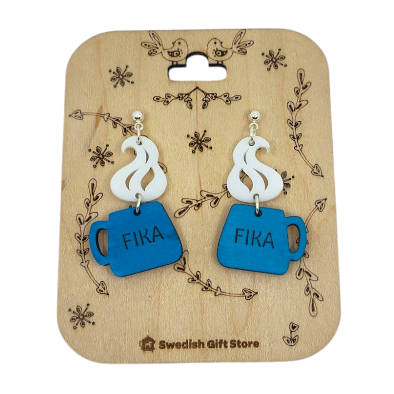 The Handmade Fika Coffee Cup Earrings showcase blue acrylic, cup-shaped designs with FIKA and steam details, beautifully displayed on a decorative wooden card.