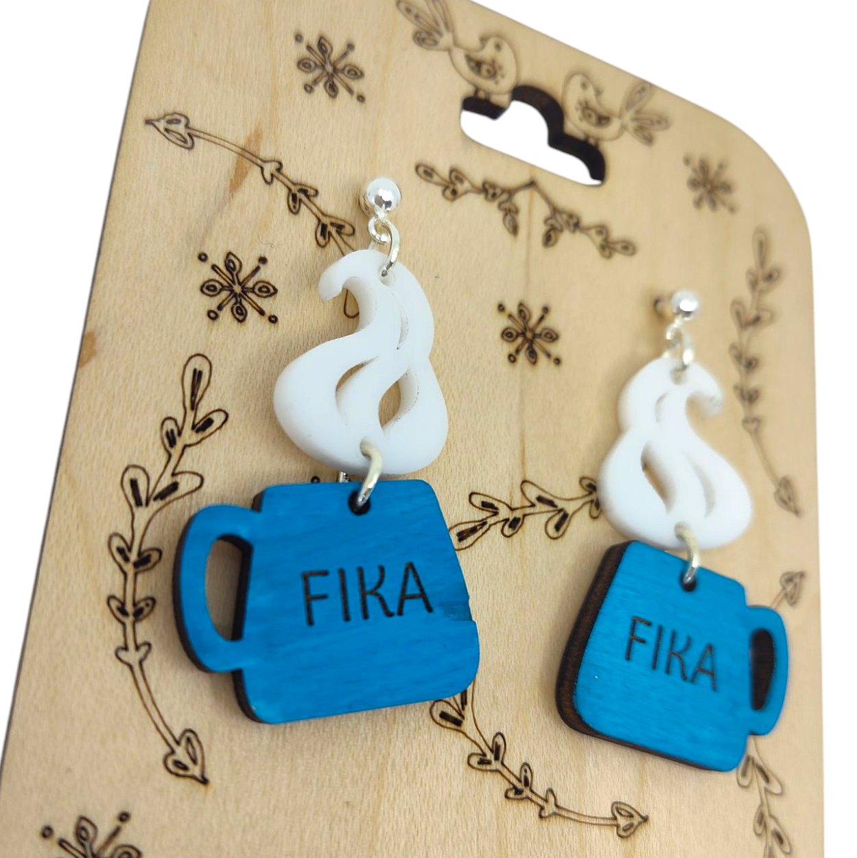 Handmade acrylic and wood earrings shaped like blue coffee cups with FIKA on them, featuring delicate white steam designs, displayed on a wooden card with floral embellishments.