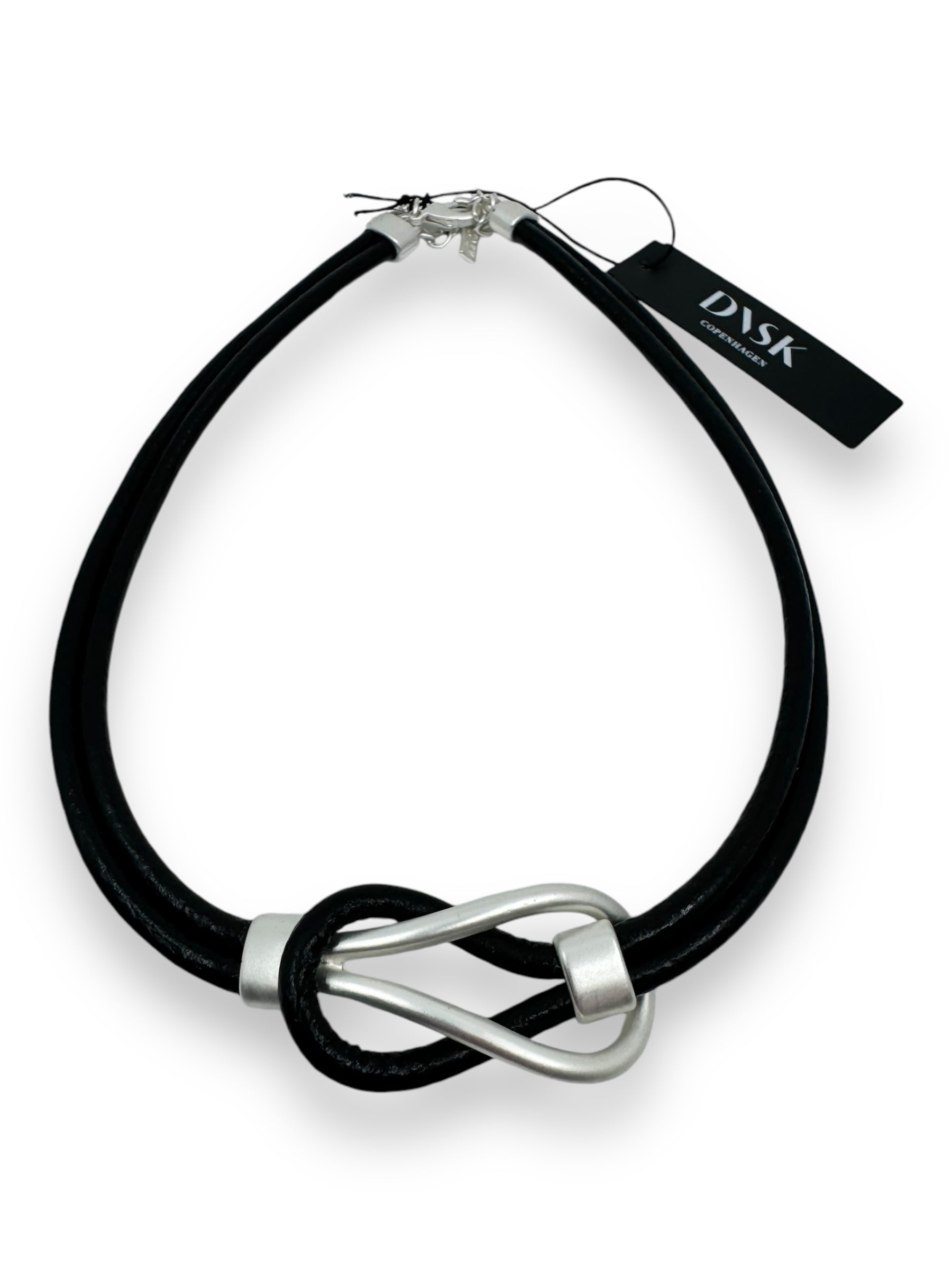 The "Togetherness Leather" necklace features a silver metallic infinity-shaped pendant with Nordic design on a black leather band. It comes with an adjustable clasp and a small black tag labeled DASK, ensuring both style and versatility.