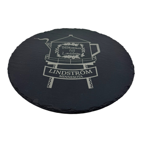 A 9.75 round black slate trivet features the Lindstrom Coffee Pot Water Tower, engraved with a teapot, floral motif, and Välkommen till Lindström, honoring this beloved Minnesota landmark.