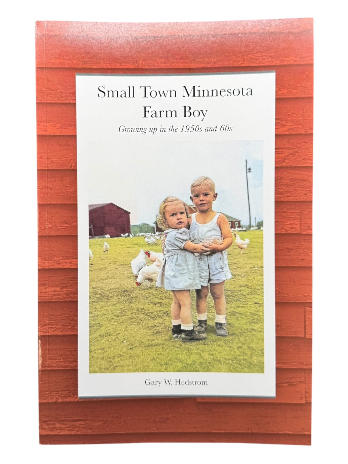 The cover of Small Town Minnesota Farm Boy – Memoir by Gary W. Hedstrom (Paperback, 274 Pages) shows two children on grass with chickens and a red barn in the background, reflecting rural mid-century Minnesota.