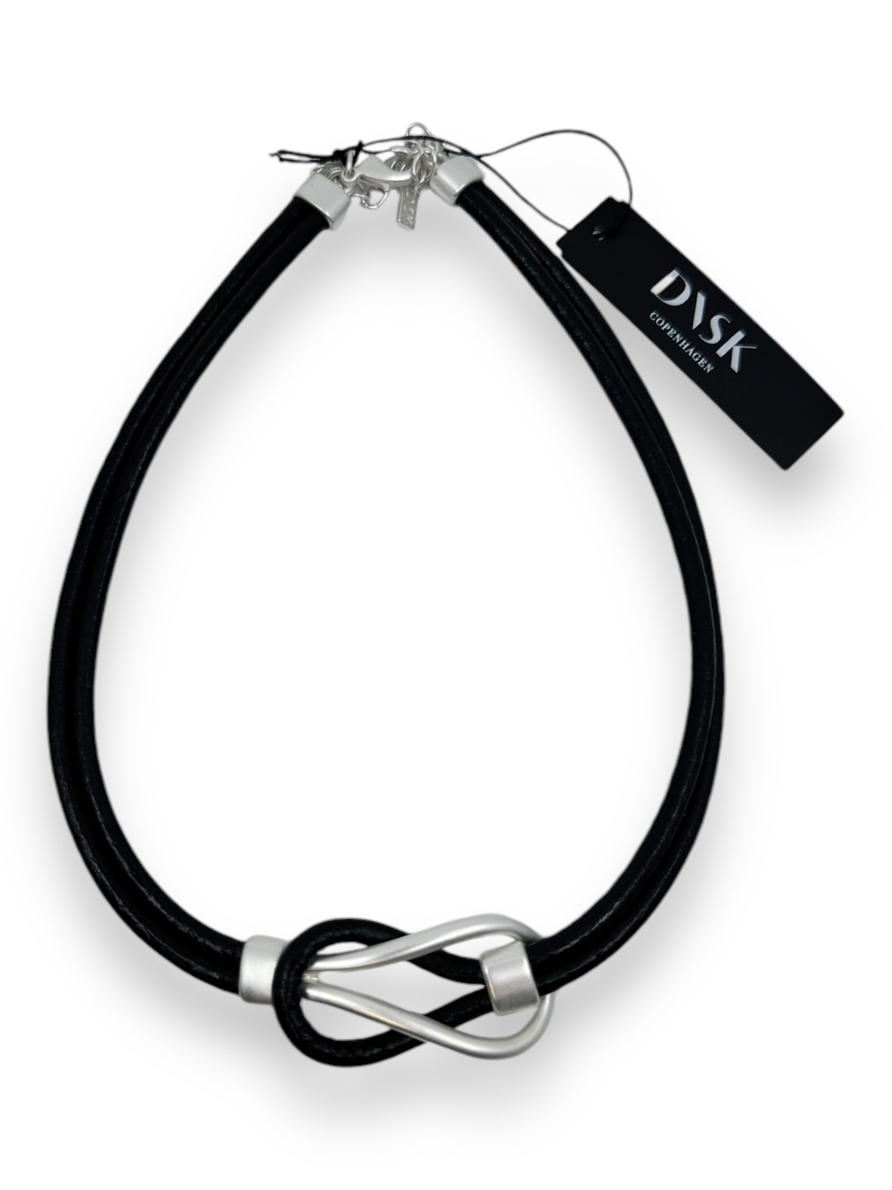 The Necklace: Togetherness Leather is a black leather necklace adorned with a silver infinity knot pendant, crafted with high-quality brass. It includes a black tag labeled DVSK Copenhagen and exemplifies Nordic design elegance, featuring an adjustable clasp for the perfect fit.