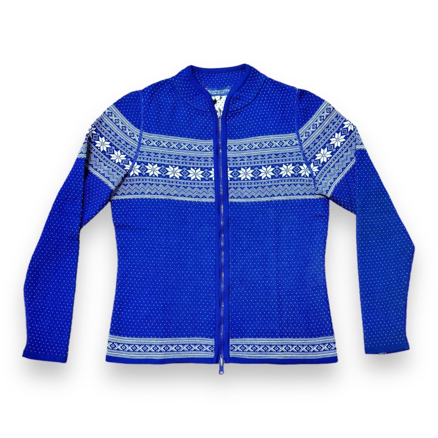 The Sweater: Cardigan Setesdal Royal Blue is a blue zip-up cardigan made from soft lamb's wool, adorned with white Nordic snowflake and geometric patterns across the chest and sleeves, adding a hint of Norwegian design.