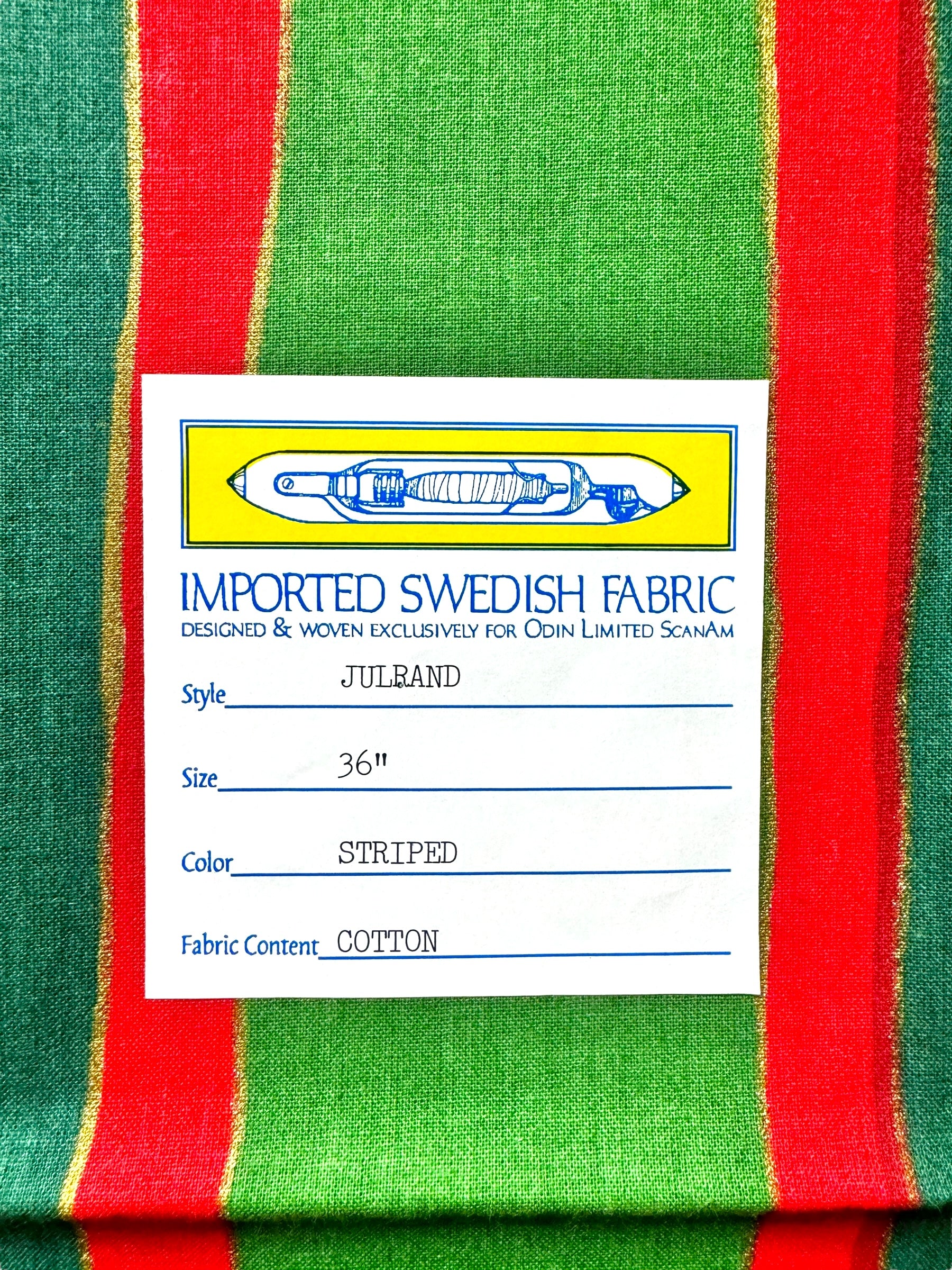 Close-up of the imported Swedish fabric label from the Runner: Julrand Holiday Runner, featuring a cotton table runner with a festive green and red stripe pattern. The label details include size (36), color (striped), and luxurious cotton fabric content.