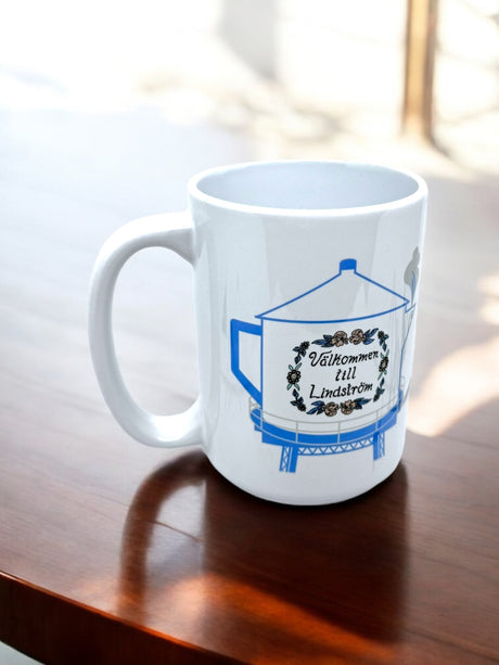 The 15oz Valkommen to Lindström Swedish Coffee Pot Water Tower Mug features the Lindström Water Tower, “Välkommen till Lindström” text, and a floral wreath—perfect for celebrating Americas Little Sweden.