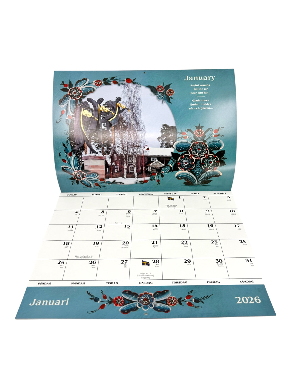 The Calendar: Sweden 2026 Wall Calendar – Visions & Verse of a Land and Its People is open to January, displaying floral patterns, a winter village scene, Swedish weekdays, and dates—showcasing the charm of Swedish culture.