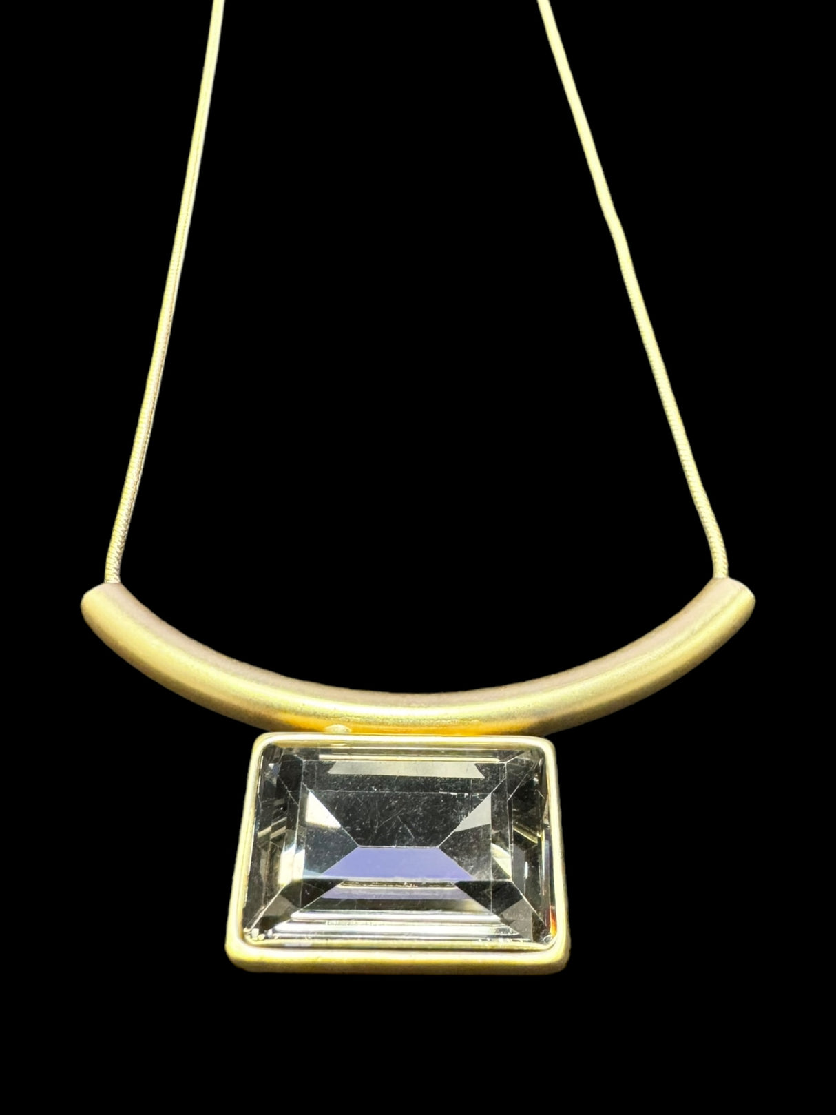 The Necklace: Audrey Black Diamond Gold Plated by DANSK Copenhagen exudes timeless elegance with its minimalist design featuring a curved gold bar, large rectangular gem, and thin chain set against a black backdrop. Its simplicity makes it a versatile accessory for any occasion.