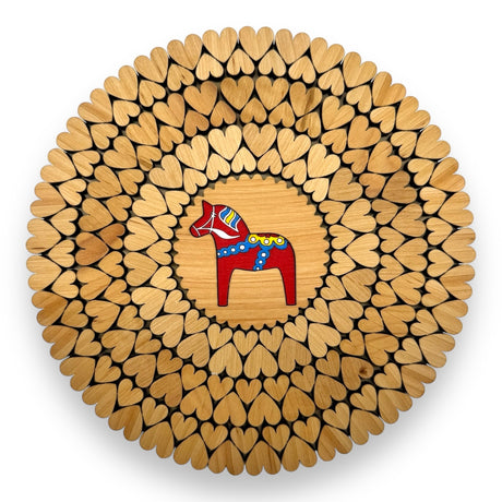 An 8" alder wood trivet features a red Swedish Dala horse at the center, surrounded by laser-cut heart shapes. This handmade piece from Estonia is displayed from above on a white background.