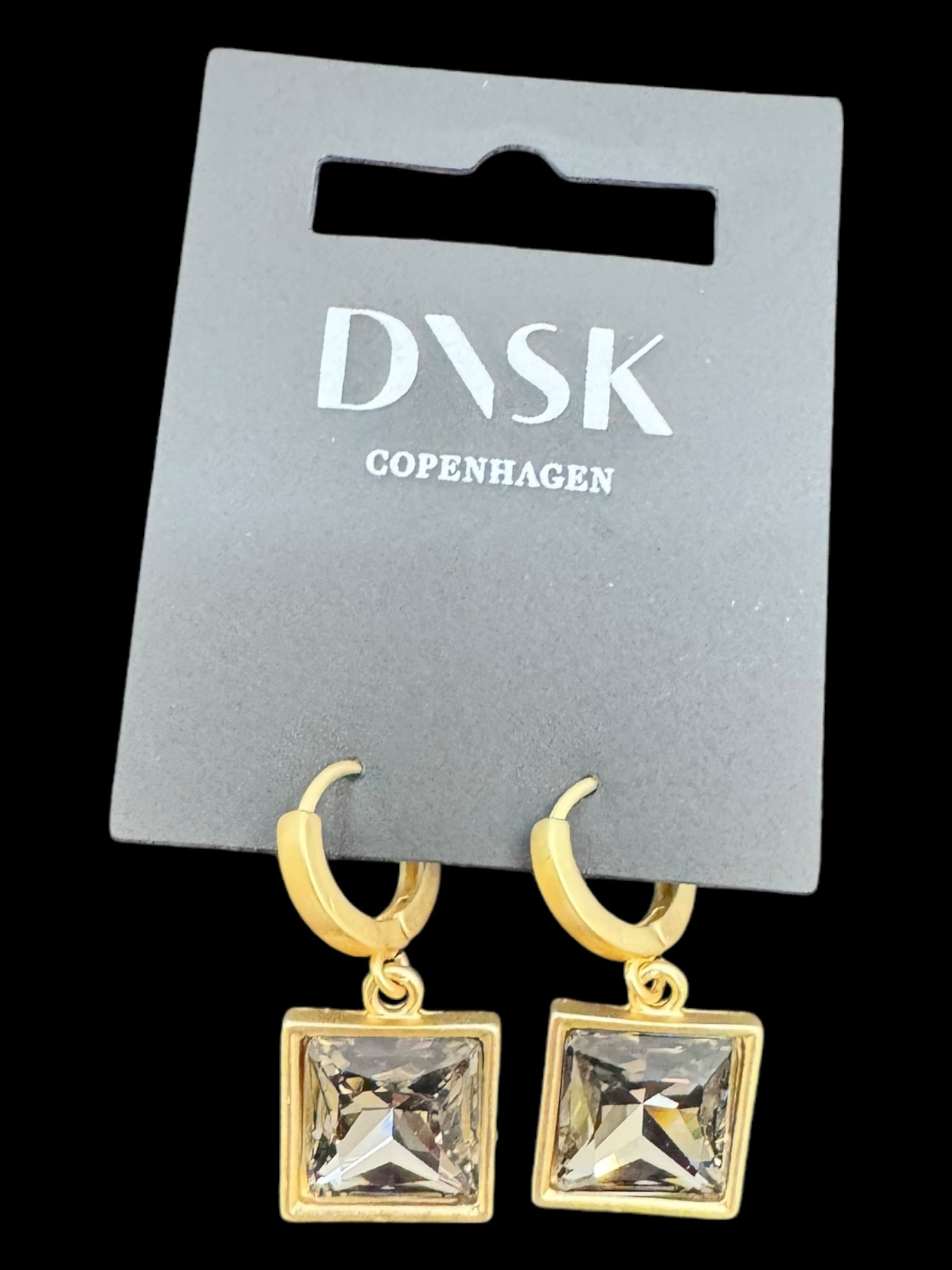 The Audrey Black Diamond Gold Plated earrings, featuring gold hoops with square clear gemstones, are presented on a gray DVSN Copenhagen display card, showcasing high-quality brass craftsmanship.