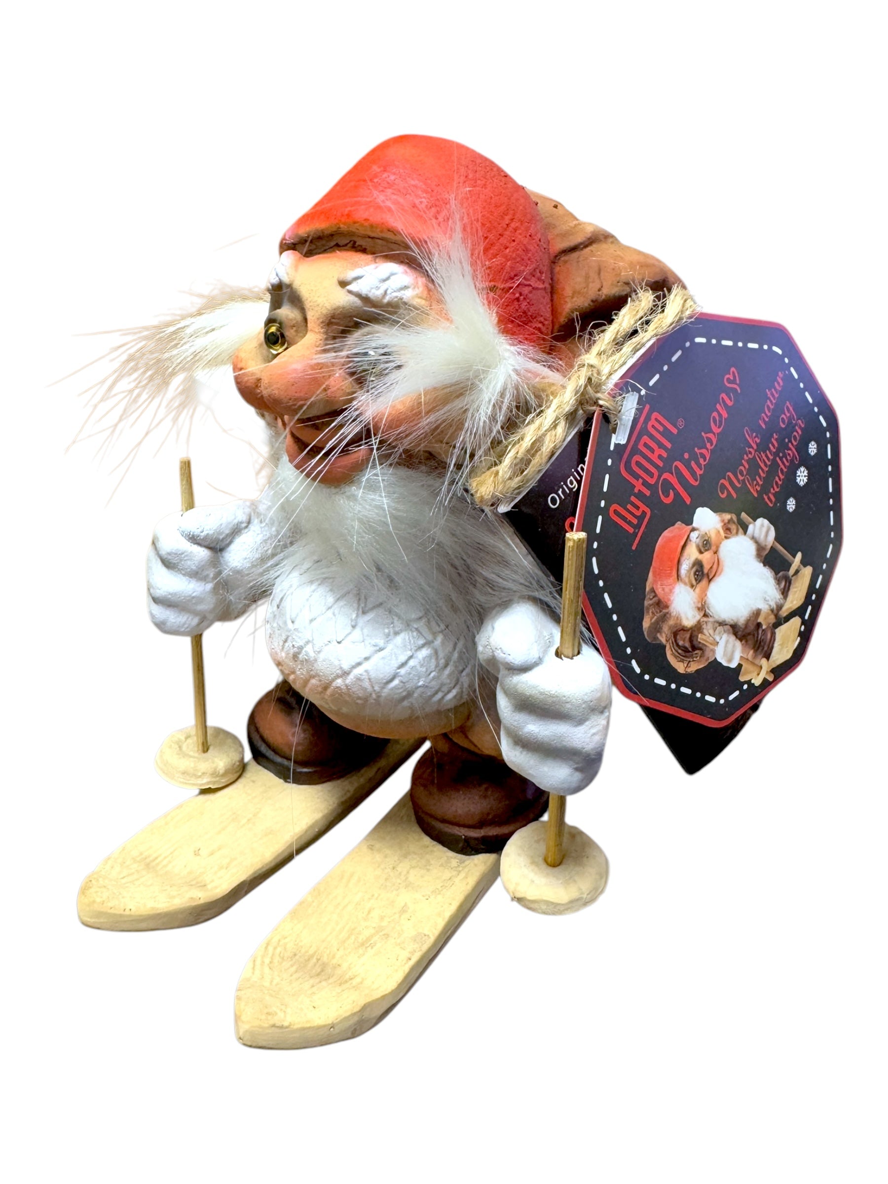 The NyForm - Nissen on Skis with Backpack is a charming collector's item crafted from wood, featuring a gnome wearing a red hat and white sweater as it skis. The attached tag showcases an illustration of another gnome along with text that embodies the essence of Norwegian design.