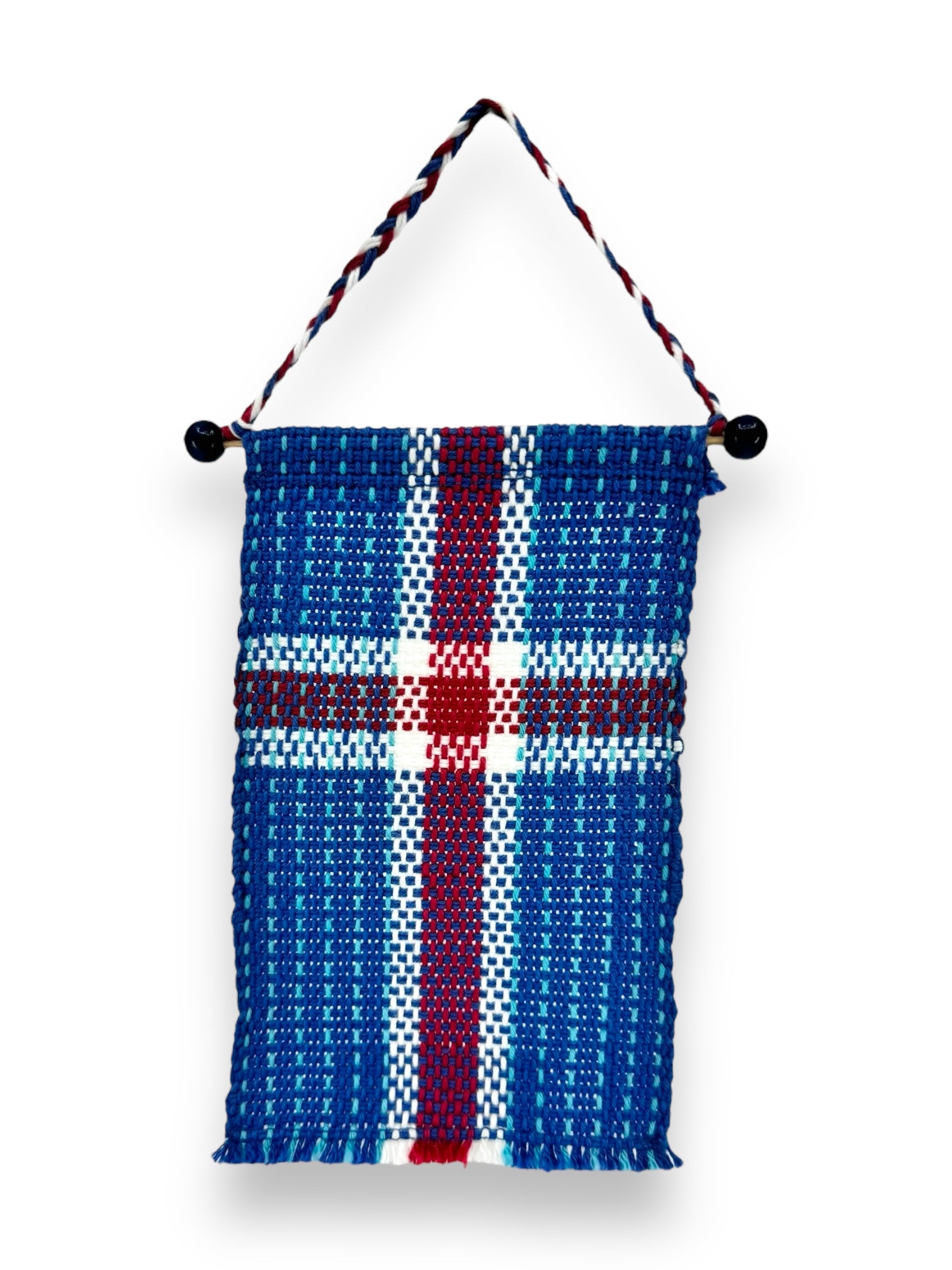 Introducing the "Flag: Small Iceland Flag on Hanger (6.5"x4")," this hand-woven wall hanging showcases a Nordic cross design in blue, white, and red hues echoing the Icelandic Flag. It is crafted from cotton string and hangs gracefully by a braided loop with black beads embellishing each side.