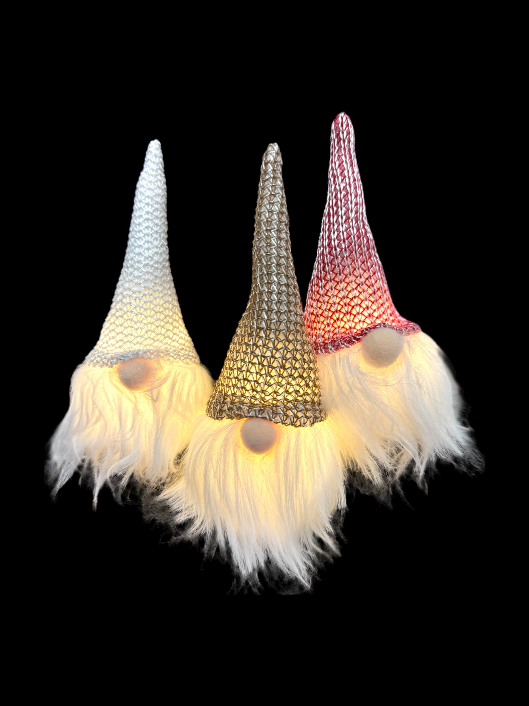 Introducing the "Ornament: Light Up Tomte Gnome," a charming set of three decorative gnomes featuring light-up noses and long, white beards. Each gnome is powered by included battery-operated A76 (LR44) cells and is adorned with tall, knitted hats available in white, silver, and red against a black background.